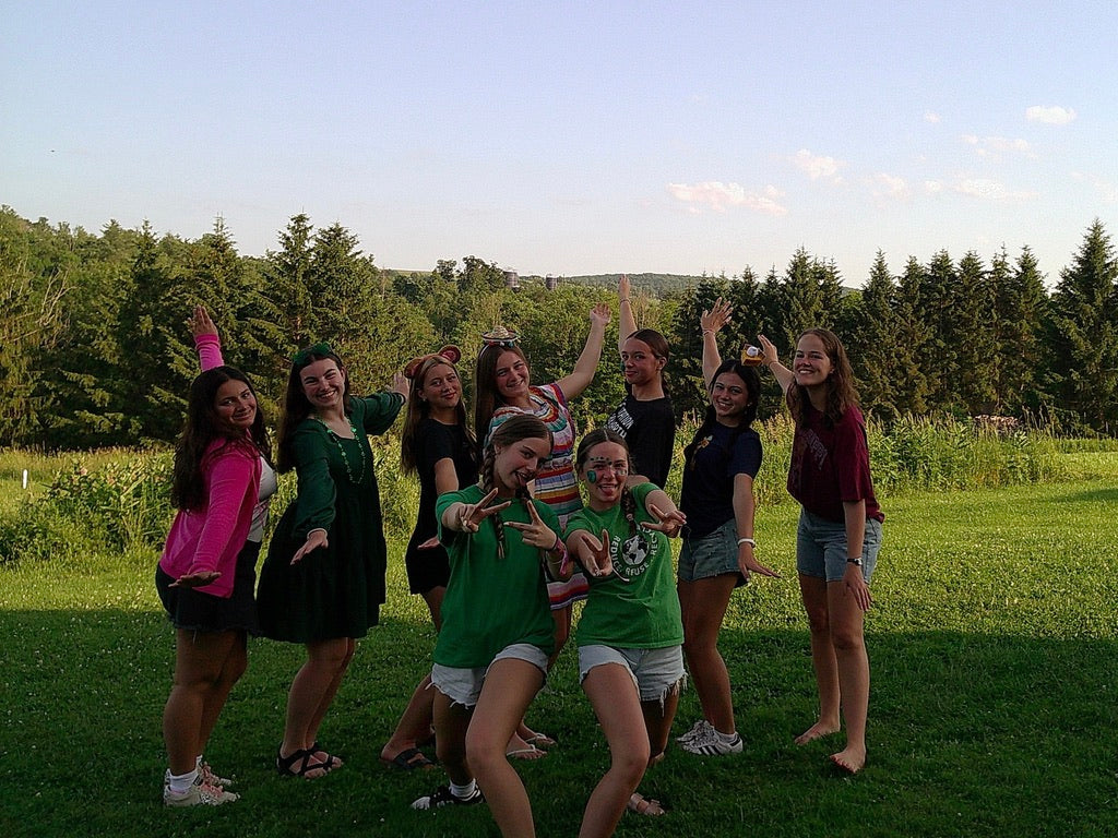 9 ladies posing in grass near the edge of a forest.